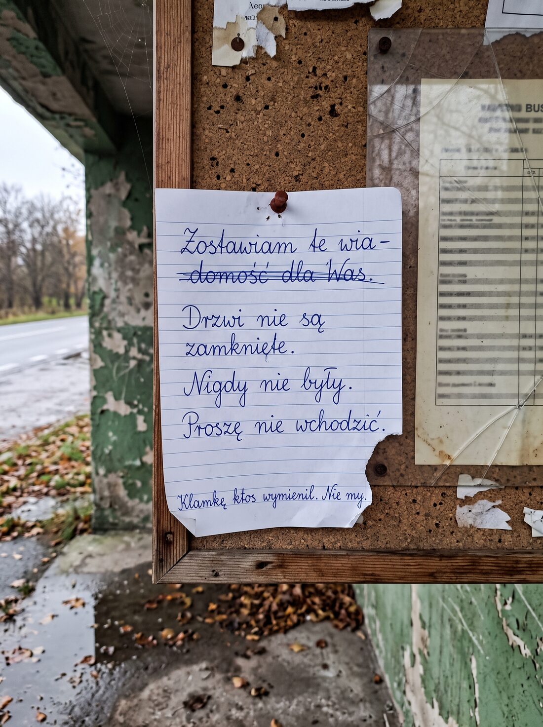 Kartka w linie z odręcznym tekstem, przypięta pinezką do korkowej tablicy ogłoszeń w betonowej wiacie przystankowej.